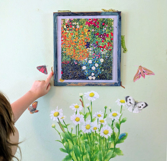 A framed colourful floral artwork hangs on a pale green wall, with a person's hand pointing at it. The scene also includes decals of daisies, butterflies, and caterpillars on the wall.
