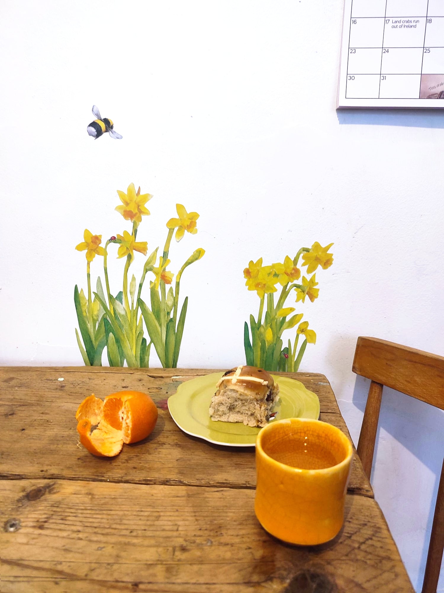 table with hot cross bun on a plate, and orange, a drink, daffodils on a white wall.