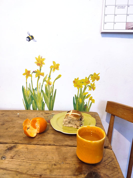 table with hot cross bun on a plate, and orange, a drink, daffodils on a white wall.