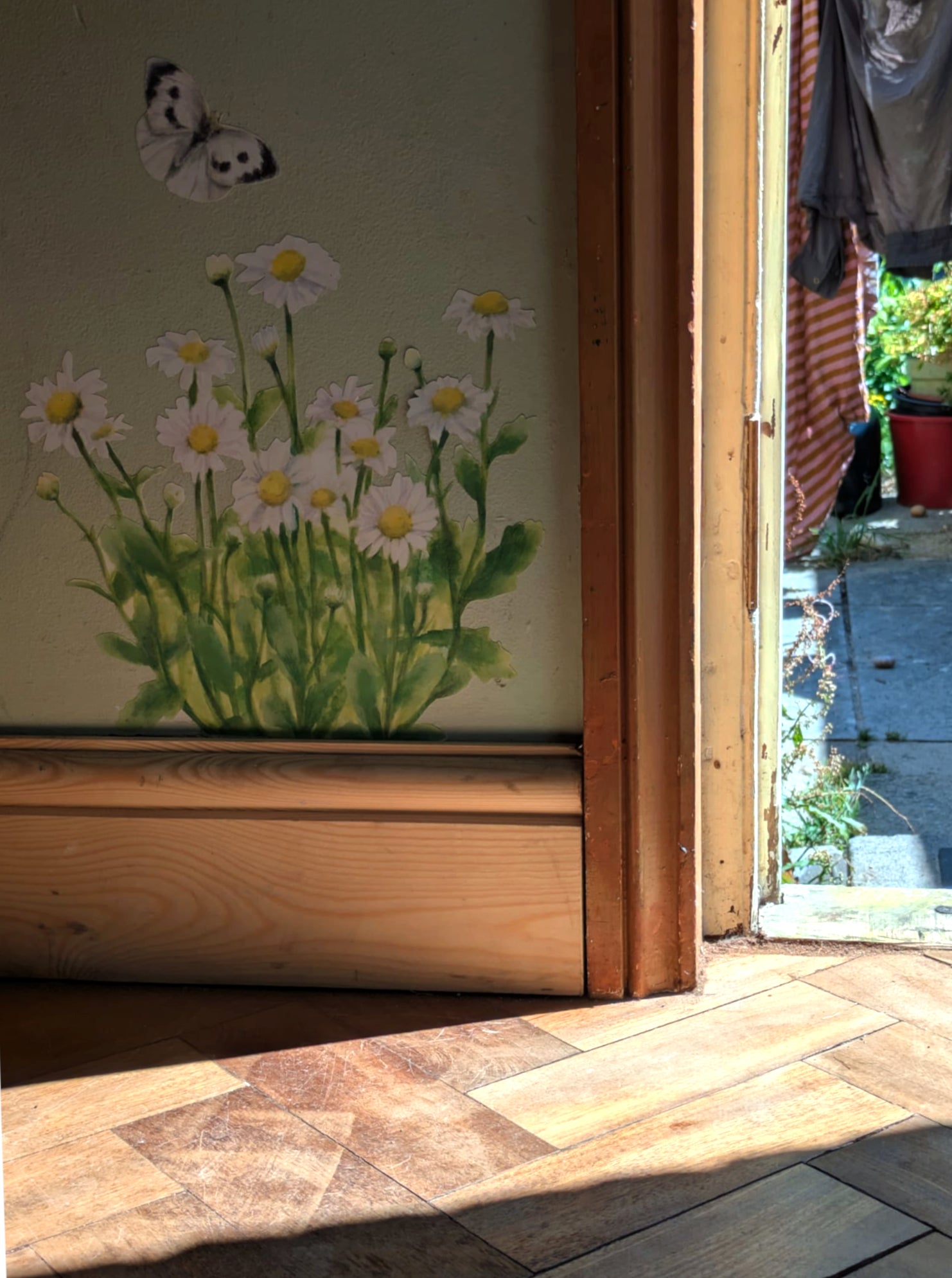 A painted wall mural featuring white daisies with yellow centers and green stems, along with a small butterfly, shown in a sunlit room next to a wooden doorway and tiled floor.