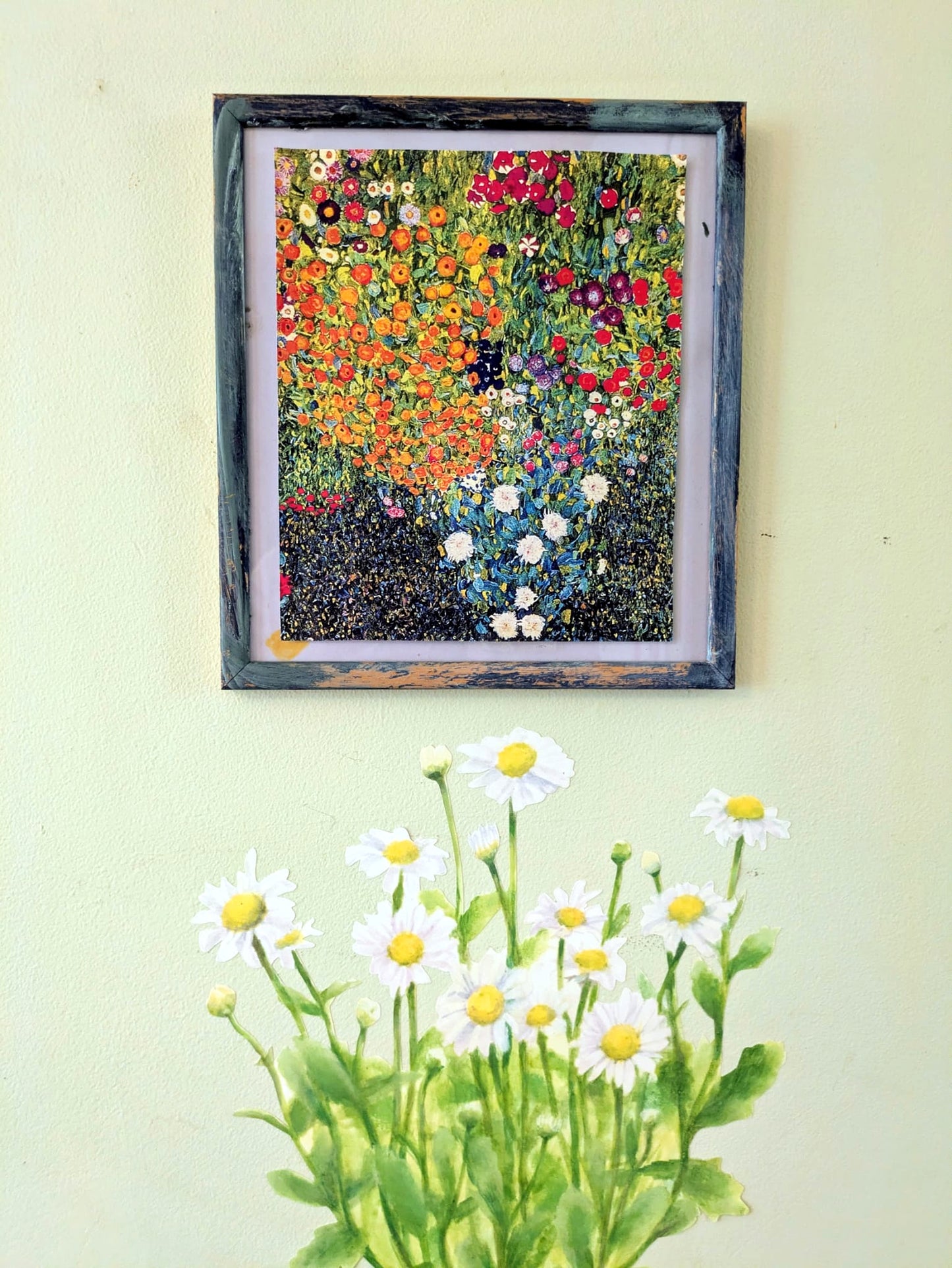 A framed floral art print hangs on a pale yellow wall above a decal of white daisies with yellow centers and green stems. 