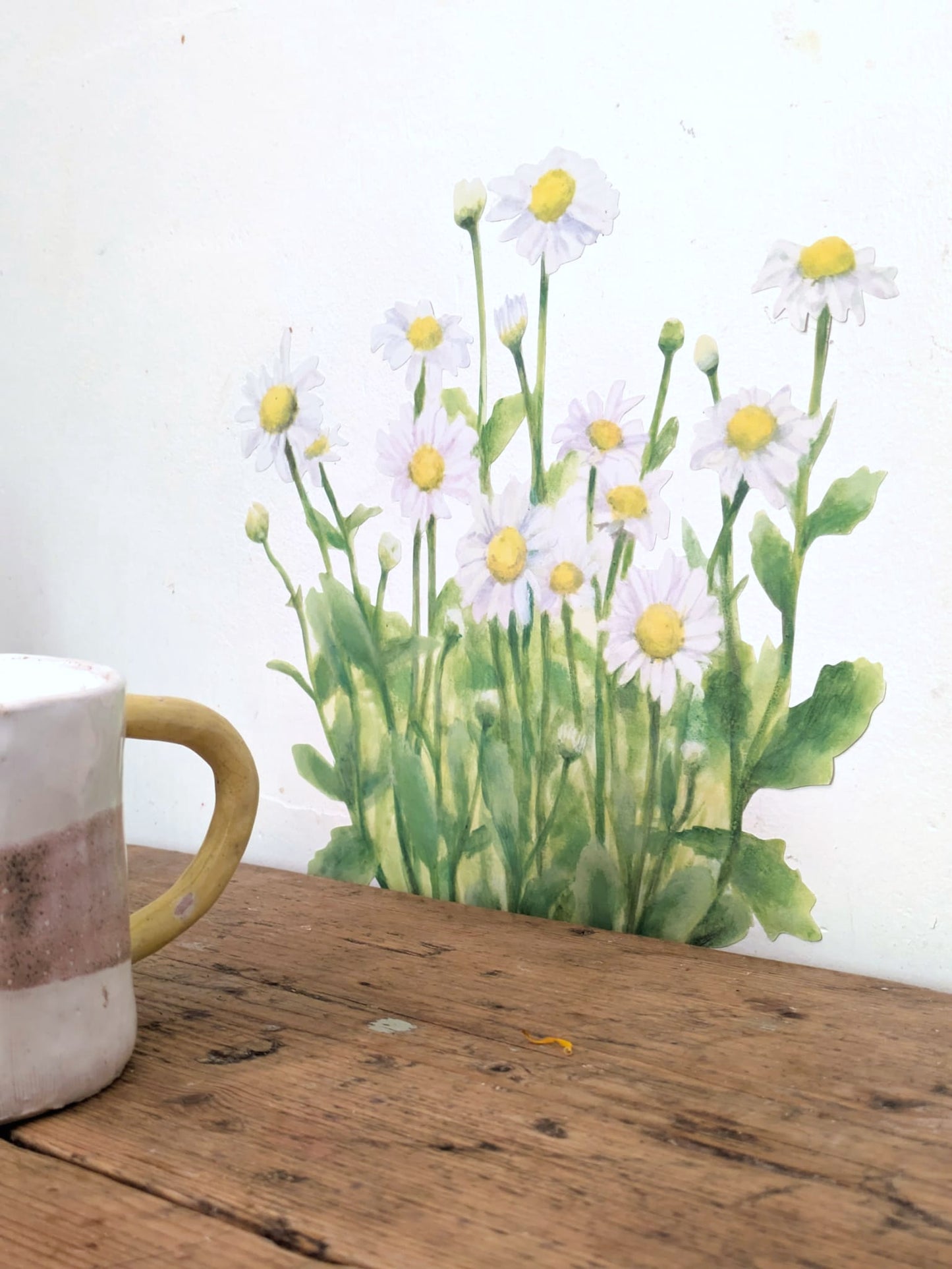A watercolor-style decal of white daisies with yellow centers and green stems, shown against a white wall above a rustic wooden tabletop. A partial ceramic mug is visible in the lower left corner.