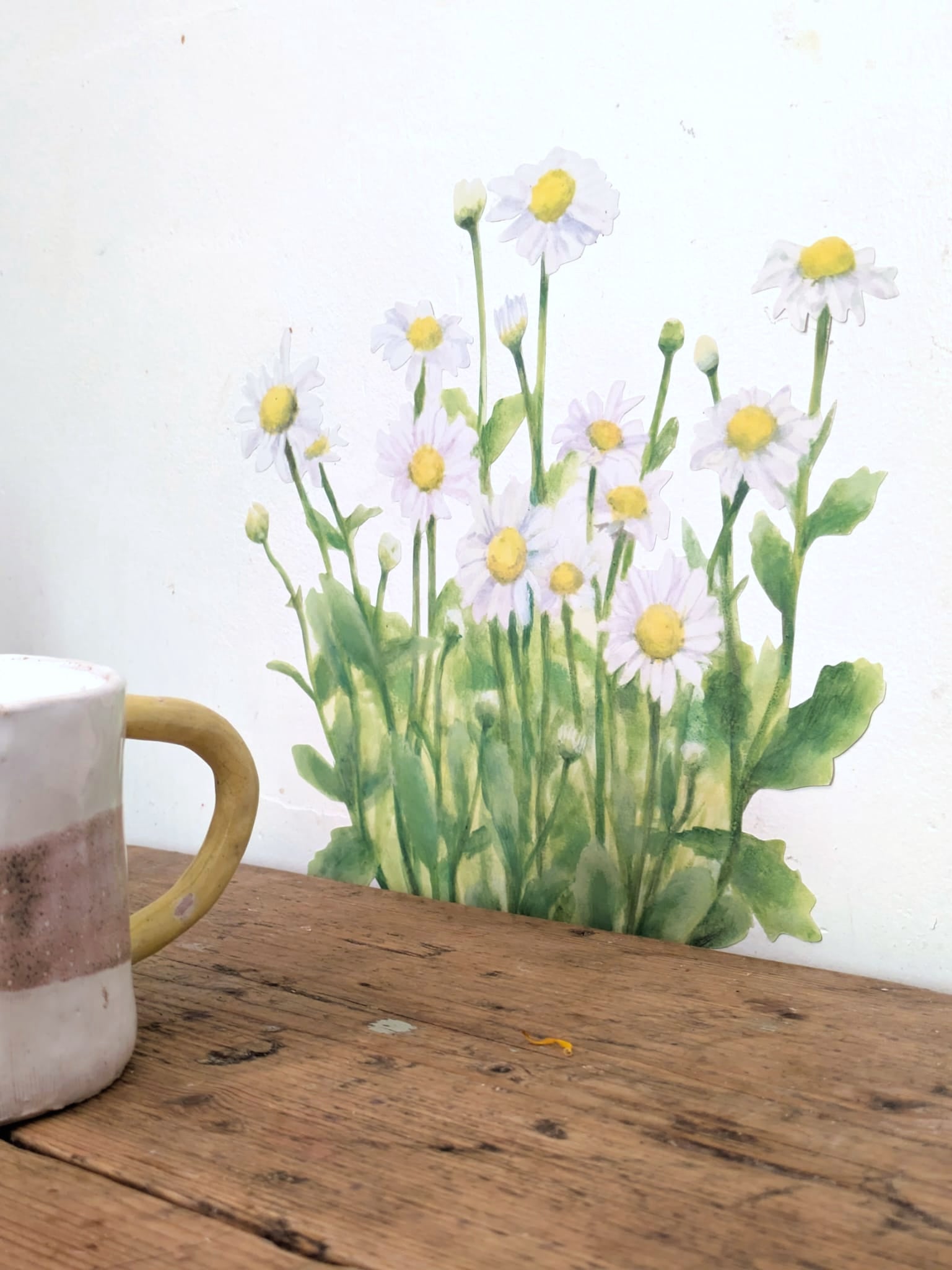 A watercolor-style decal of white daisies with yellow centers and green stems, shown against a white wall above a rustic wooden tabletop. A partial ceramic mug is visible in the lower left corner.