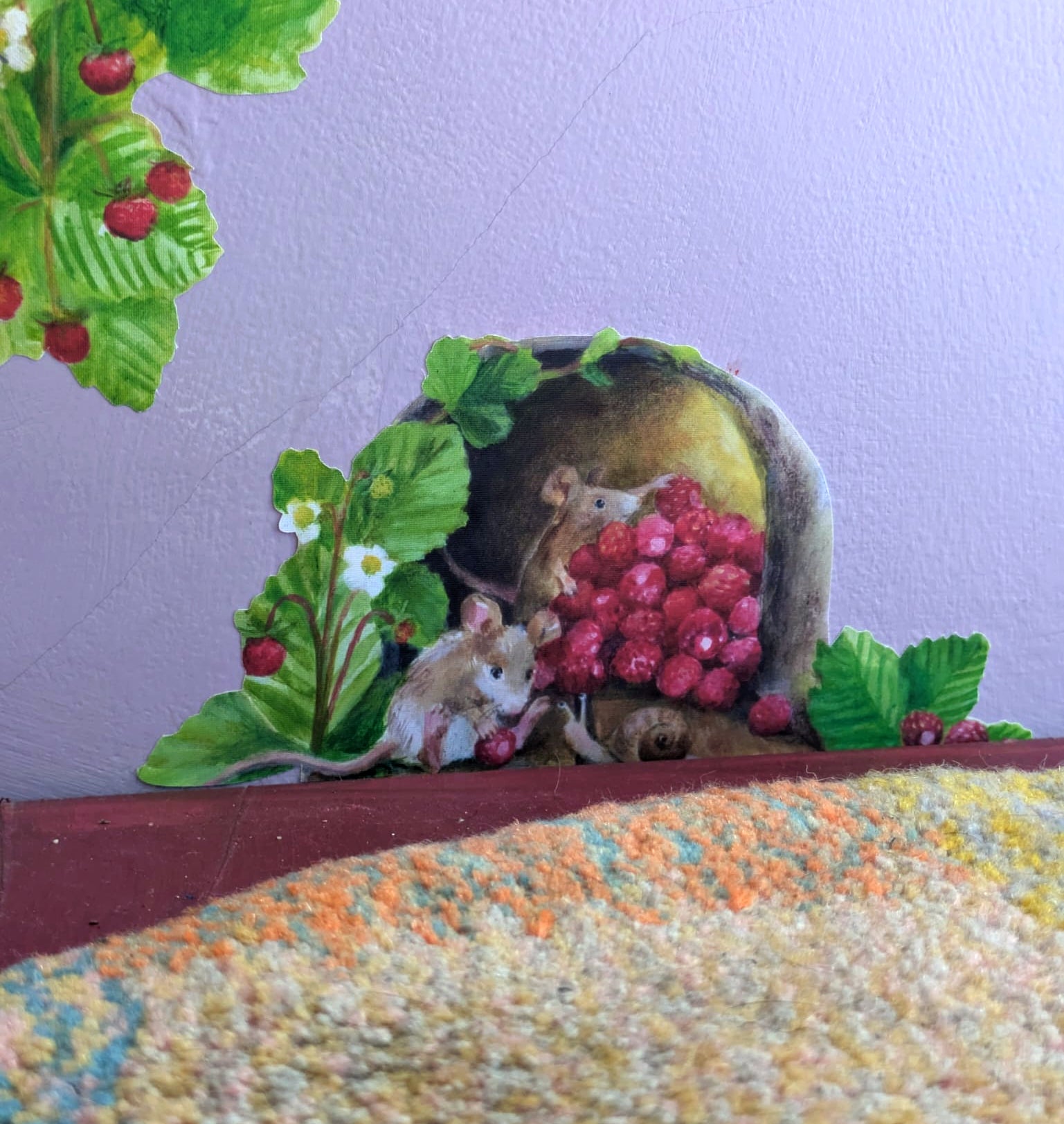 A decorative wall decal featuring a painted scene of small brown mice among strawberry leaves, red berries, and a yellow fruit on a pale lavender background, with a textured multicolor rug in the foreground.