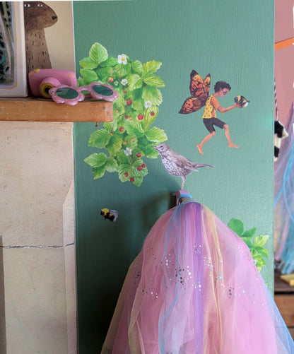 A whimsical fairy-themed nursery wall decoration featuring a painted fairy, bird, bee, and strawberry plant on a green panel, with a pink and purple tulle dress hanging below.