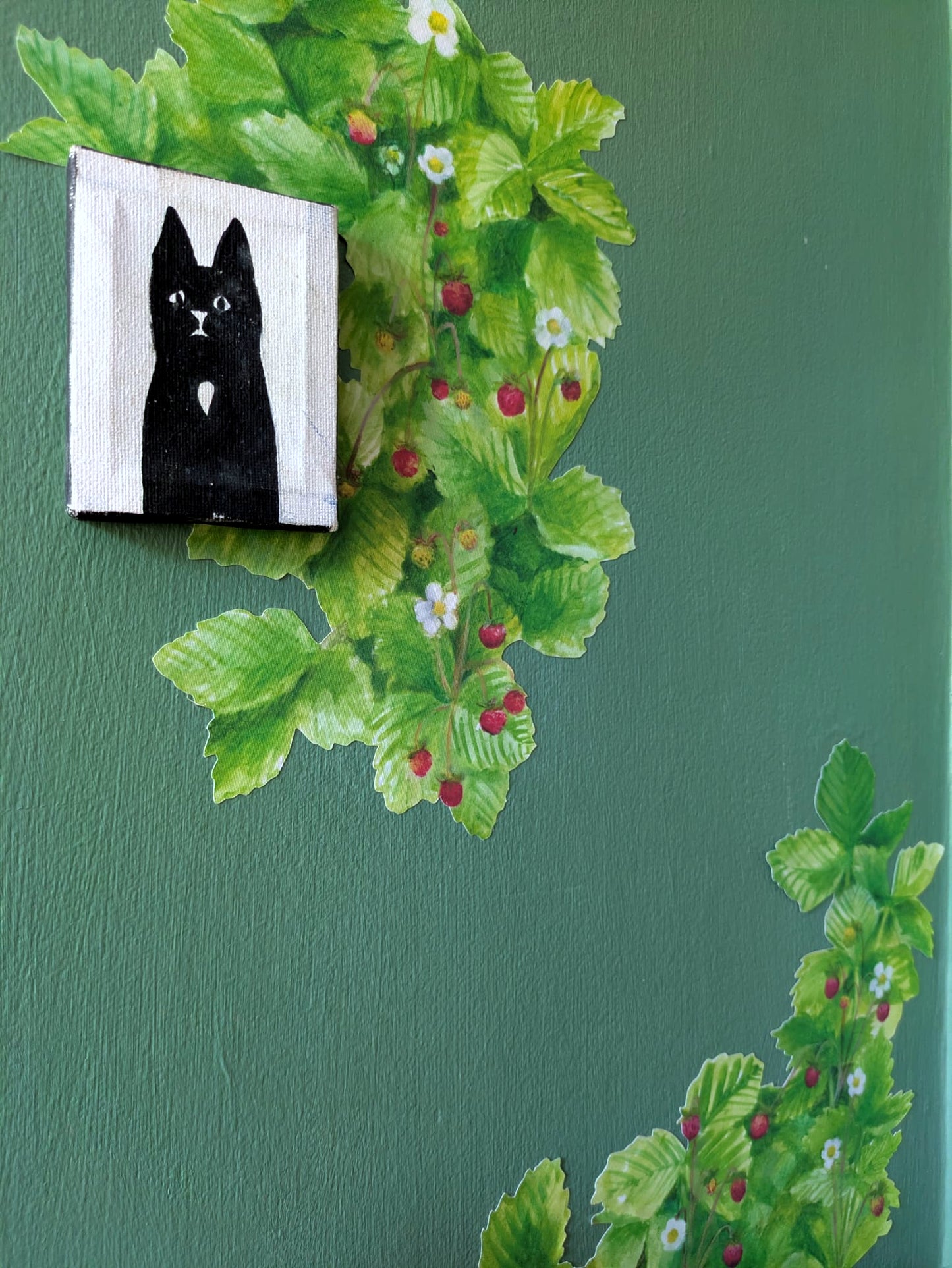 A square canvas print featuring a black cat on a white background, mounted on a textured dark green wall with a wild strawberry decal flowing down behind it.