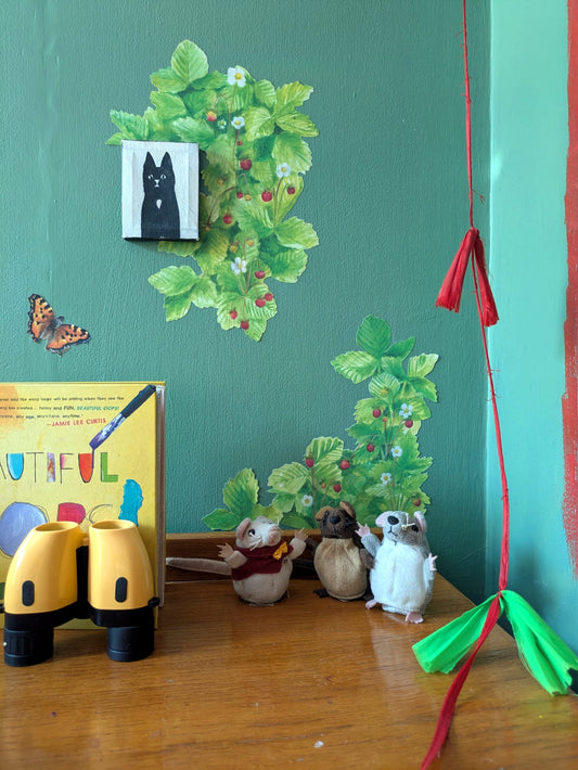 A styled corner of a room with a teal wall, featuring a black cat portrait print, wild strawberry wall decals, a butterfly sticker, a yellow children's book, yellow toy binoculars, three small mouse figurines, and hanging red and green ribbon decorations.