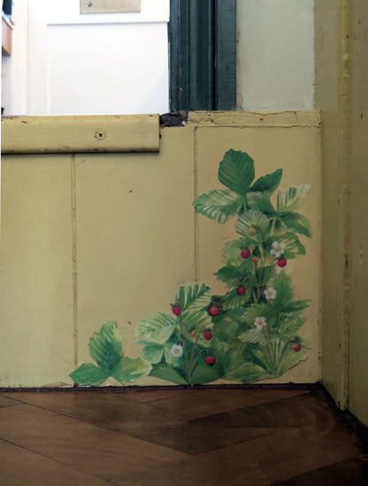 A painted corner of a cream-colored wall with a green strawberry plant motif, above a dark wood floor.
